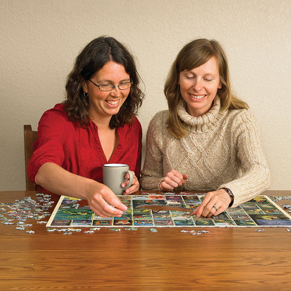 Two women enjoying a Nancy Drew jigsaw puzzle by Cobble Hill, working together on a wooden table with various puzzle pieces scattered around. One woman holds a gray mug while the other focuses on placing a piece.