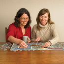Two women enjoying a vintage American postcards jigsaw puzzle. One woman in a red shirt is placing a puzzle piece while the other, wearing a cozy beige sweater, smiles. A mug is on the table with scattered puzzle pieces around them.