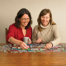 Two women enjoying a fun afternoon putting together a colorful Sugar Skull Jigsaw Puzzle called Sugar Skull Cookies by Cobble Hill. One woman, in a red shirt, is placing a puzzle piece while holding a mug, and the other, in a beige sweater, is smiling and reaching for a piece. The table is covered with scattered puzzle pieces and the partially completed puzzle features vibrant sugar skull designs.