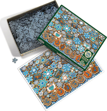The image features the Cobble Hill Hanukkah Cookies jigsaw puzzle. The puzzle box is open and shows a colorful design with blue, gold, and white motifs resembling Hanukkah cookies, stars, dreidels, and menorahs. Scattered puzzle pieces are visible within the box, creating a festive holiday scene.