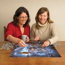 Two women engaged in assembling the Dragonforge jigsaw puzzle by Cobble Hill. One has a coffee cup, and vibrant puzzle pieces are scattered around them on a wooden table as they focus on fitting pieces together.