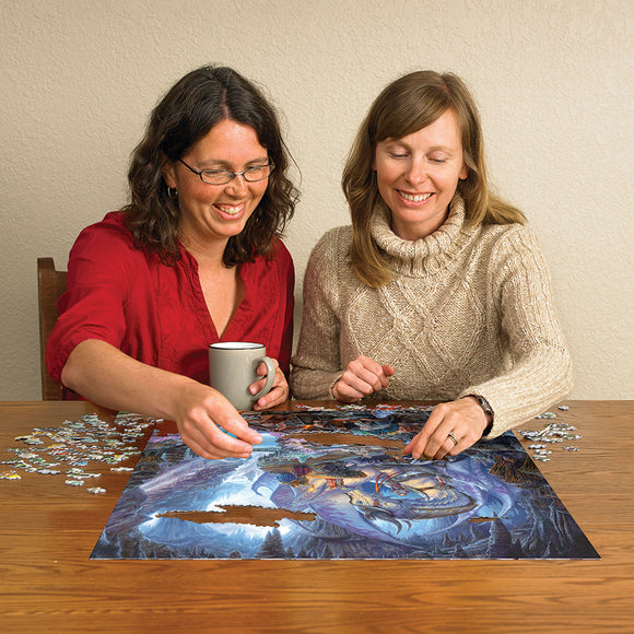 Two women engaged in assembling the Dragonforge jigsaw puzzle by Cobble Hill. One has a coffee cup, and vibrant puzzle pieces are scattered around them on a wooden table as they focus on fitting pieces together.