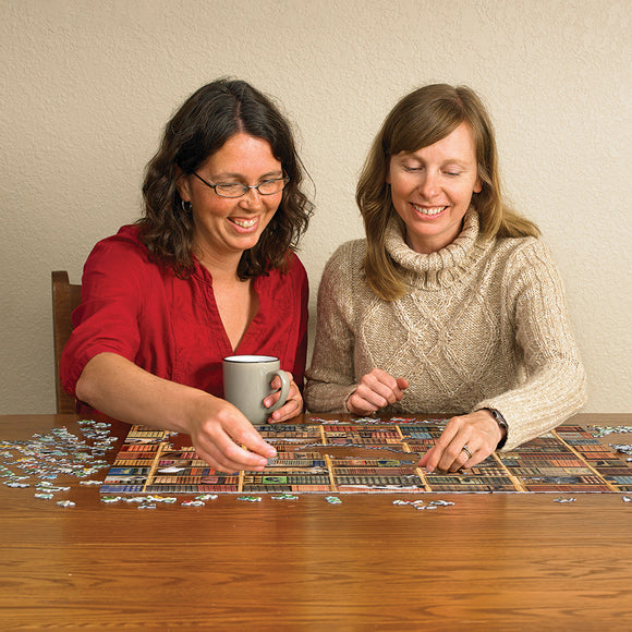 Two women enjoying The Cat Library jigsaw puzzle by Cobble Hill. One woman is holding a grey mug while picking up a piece. The table is covered with puzzle pieces and the nearly completed puzzle, showcasing colorful cat-themed artwork.
