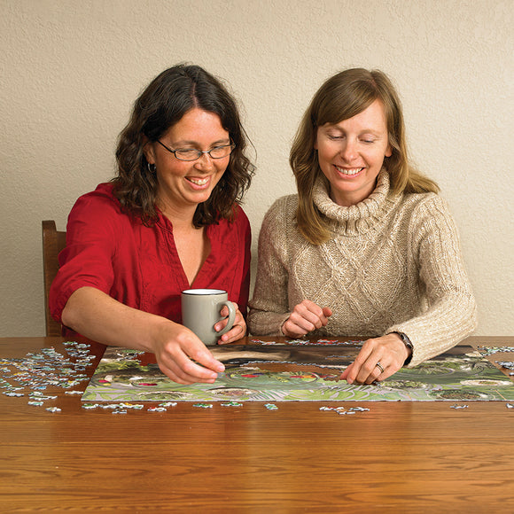 Two women enjoying a jigsaw puzzle titled 'Terrarium Cat' by Cobble Hill, sitting at a wooden table surrounded by puzzle pieces. One woman holds a gray mug while they both smile and piece together the vibrant puzzle featuring a cat among lush greenery.