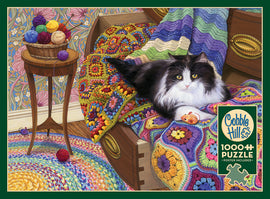 A 1000-piece jigsaw puzzle titled 'Comfy Cat' by Cobble Hill, featuring a cozy scene with a black and white cat lounging on a colorful crocheted blanket. Surrounding the cat are vibrant yarn balls in a basket, with intricate floral wallpaper in the background, creating a warm and inviting atmosphere.