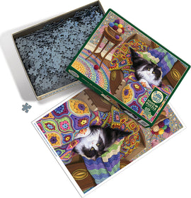 The Comfy Cat jigsaw puzzle by Cobble Hill features a charming illustration of a cat lounging on a colorful crocheted blanket, surrounded by vibrant decor and a basket of yarn. The puzzle pieces are shown in an open box, highlighting their intricate shapes.