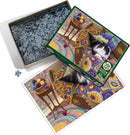 Cobble Hill's Comfy Cat jigsaw puzzle featuring a cozy black and white cat resting on a colorful crochet blanket. The puzzle box is partially open, revealing intricately cut puzzle pieces inside. An image of the completed puzzle is displayed beside the box, showcasing vibrant colors and detailed patterns, perfect for puzzle enthusiasts.