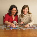 Two women enjoying their time together as they work on the Comfy Cat jigsaw puzzle by Cobble Hill. One woman holds a grey mug while carefully placing a piece, and colorful puzzle pieces are scattered around a wooden table.