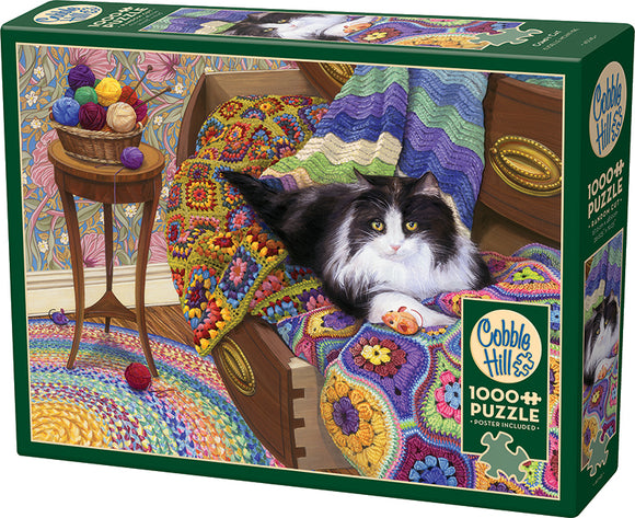 Comfy Cat jigsaw puzzle by Cobble Hill features a cozy scene with a fluffy black and white cat lounging on a colorful, crocheted blanket surrounded by vibrant yarn balls. The puzzle comes with 1000 pieces and includes a poster for reference.