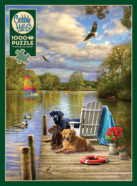 A 1000-piece jigsaw puzzle titled 'Dog Day Afternoon' by Cobble Hill, featuring a serene lakeside scene with two dogs lounging on a dock. In the background, a colorful sailboat is seen on the water and trees with autumn foliage line the shore. A white adirondack chair sits beside a small table adorned with flowers, adding a welcoming touch to the tranquil setting. Birds are flying and swimming nearby, enhancing the peaceful outdoor atmosphere.