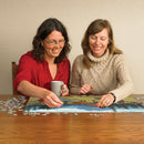 Two women joyfully working together on the Horse Meadow jigsaw puzzle by Cobble Hill. One is holding a coffee mug while placing a puzzle piece, and the other watches intently, both enjoying this creative and relaxing activity. The wooden table is scattered with colorful puzzle pieces, and the partially completed puzzle showcases a scenic meadow.