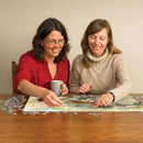 Two women engaged in assembling the Hidden Nest jigsaw puzzle by Cobble Hill. One woman is holding a mug while placing a puzzle piece, with completed sections visible on the wooden table surrounded by scattered pieces.