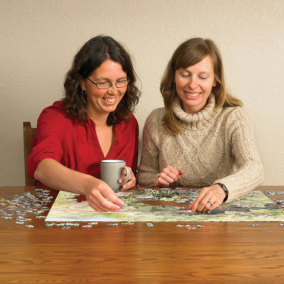 Two women engaged in assembling the Hidden Nest jigsaw puzzle by Cobble Hill. One woman is holding a mug while placing a puzzle piece, with completed sections visible on the wooden table surrounded by scattered pieces.