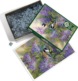 A colorful jigsaw puzzle titled 'Chickadees and Lilacs' by Cobble Hill. The box contains numerous intricately shaped pieces inside, alongside an image of the completed puzzle featuring vibrant lilacs and charming chickadee birds.