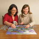 Two women seated at a wooden table assembling a jigsaw puzzle titled 'Chickadees and Lilacs' from Cobble Hill. One woman, wearing a red shirt and glasses, smiles as she places a puzzle piece, while the other, in a cozy beige sweater, observes closely with a happy expression. A steaming cup is beside the first woman, and colorful puzzle pieces are scattered around them, creating a warm, inviting atmosphere of togetherness and creativity.