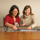 Two women engaged in solving the 'Mountain Thunder' jigsaw puzzle by Cobble Hill, enjoying a cozy atmosphere. One woman is placing a piece while holding a coffee mug, and the other is smiling, surrounded by scattered puzzle pieces on a wooden table.