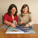 Two women enjoying a fun jigsaw puzzle activity together at a wooden table. One woman is holding a coffee mug and placing a puzzle piece, while the other assists in completing the beautiful Raven Rendezvous jigsaw puzzle by Cobble Hill, showcasing vibrant artwork of ravens amidst nature.