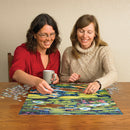 Two women enthusiastically working together on the 'Koi Pond' jigsaw puzzle by Cobble Hill. One woman is holding a coffee mug while both reach for puzzle pieces featuring vibrant water lilies and koi fish against a blue water background, showcasing a fun and interactive puzzle-solving experience.