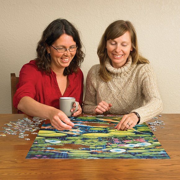 Two women enthusiastically working together on the 'Koi Pond' jigsaw puzzle by Cobble Hill. One woman is holding a coffee mug while both reach for puzzle pieces featuring vibrant water lilies and koi fish against a blue water background, showcasing a fun and interactive puzzle-solving experience.