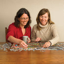 Two women sitting at a wooden table, working together on a colorful jigsaw puzzle featuring fishing lures. One woman holds a gray mug, while both women are smiling and engaged in the activity.
