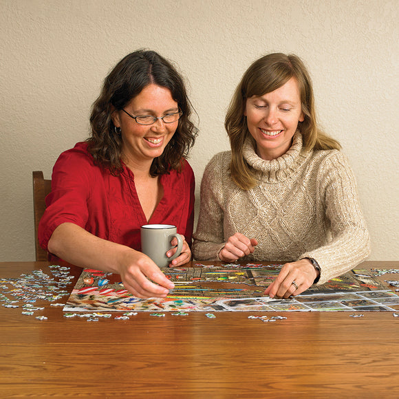 Two women sitting at a wooden table, working together on a colorful jigsaw puzzle featuring fishing lures. One woman holds a gray mug, while both women are smiling and engaged in the activity.