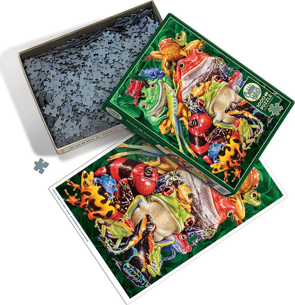 A colorful jigsaw puzzle titled 'Frog Business' by Cobble Hill. The puzzle box, open to reveal its contents, displays various vibrant frog illustrations on the lid, featuring different species in an array of colors. The puzzle pieces are arranged in the box, with a single piece set outside, on a white surface. An image of the completed puzzle is shown next to the box, emphasizing the intricate design of the frogs against a lush green background.