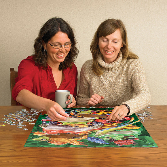 Two women engaged in assembling the 'Frog Business' jigsaw puzzle by Cobble Hill. One woman holds a coffee cup while they both work on a colorful puzzle featuring various frogs in a vibrant green setting.