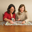 Two women sitting at a wooden table, working together on a jigsaw puzzle featuring botanical illustrations. One woman holds a gray mug while the other places a puzzle piece. Small, scattered puzzle pieces surround them.
