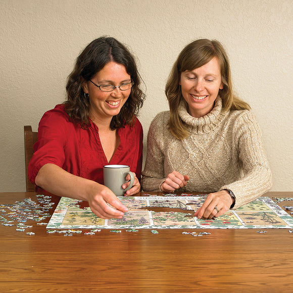 Two women sitting at a wooden table, working together on a jigsaw puzzle featuring botanical illustrations. One woman holds a gray mug while the other places a puzzle piece. Small, scattered puzzle pieces surround them.