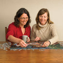 Two women smiling and assembling the 'Christmas Puppies' jigsaw puzzle from Cobble Hill. One woman holds a coffee mug while placing a puzzle piece on a festive scene featuring puppies and holiday decorations.