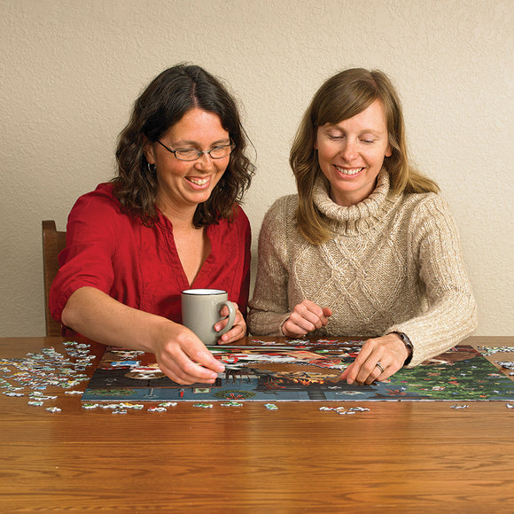 Two women smiling and assembling the 'Christmas Puppies' jigsaw puzzle from Cobble Hill. One woman holds a coffee mug while placing a puzzle piece on a festive scene featuring puppies and holiday decorations.
