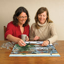 Two women enjoying a jigsaw puzzle titled 'Village Tree' by Cobble Hill. One woman holds a coffee mug while placing a puzzle piece on the table. The partially assembled puzzle features a village scene with a large green tree and cozy houses, surrounded by scattered puzzle pieces on a wooden table.