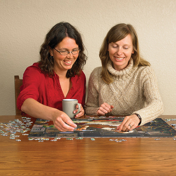 Two women smiling while assembling the Santa Painting Cars jigsaw puzzle on a wooden table. One woman holds a gray mug, while they work together on the colorful puzzle pieces scattered around them.