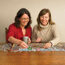 Two women are enjoying a jigsaw puzzle titled 'DoodleTown: Elves at Work' by Cobble Hill, sitting at a wooden table with scattered puzzle pieces. One woman holds a coffee mug as they both focus on the puzzle, which features colorful, whimsical scenes with elves.