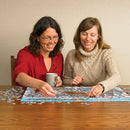 Two women engaged in assembling the DoodleTown: 12 Days of Christmas jigsaw puzzle on a wooden table, surrounded by scattered puzzle pieces. One woman is holding a gray mug while joyfully placing a puzzle piece, and the other is smiling as they collaborate on the puzzle together, creating a festive atmosphere.
