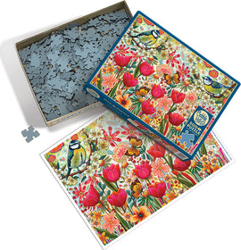The 'Shooting the Breeze' jigsaw puzzle by Cobble Hill featuring a colorful illustration of flowers, a blue tit bird, and butterflies. The box is partially opened, revealing the puzzle pieces inside and the completed image displayed beside it.