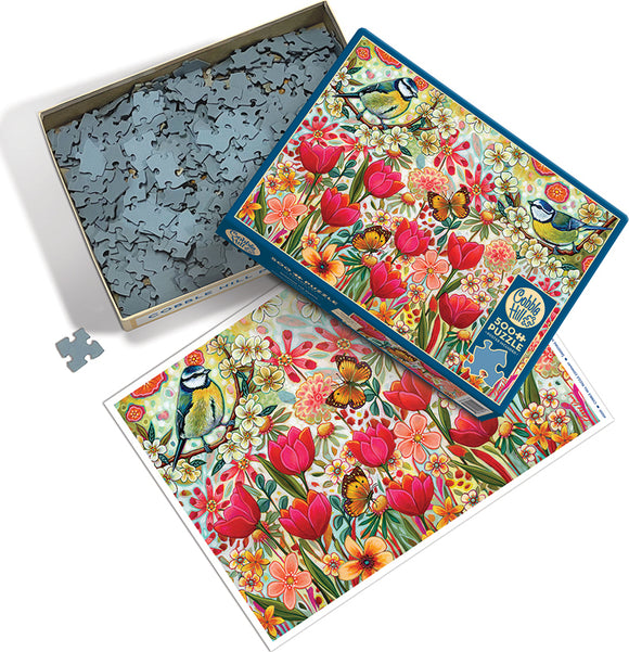 The 'Shooting the Breeze' jigsaw puzzle by Cobble Hill featuring a colorful illustration of flowers, a blue tit bird, and butterflies. The box is partially opened, revealing the puzzle pieces inside and the completed image displayed beside it.