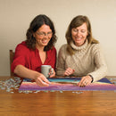 Two women enjoying a jigsaw puzzle featuring the design 'Crossings: Thunderbird and Bear' by Cobble Hill. They are sitting at a wooden table, one holding a mug, as they piece together the colorful image surrounded by scattered puzzle pieces.