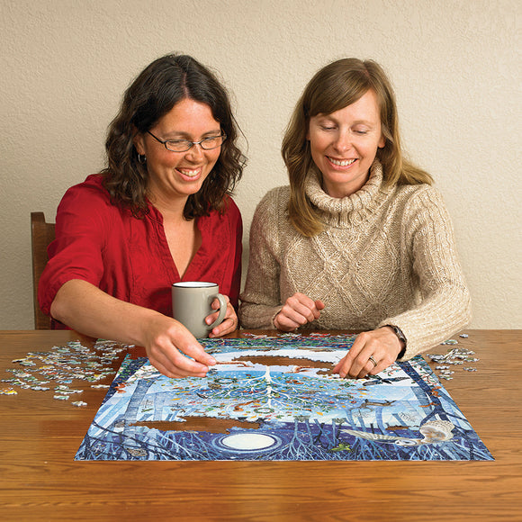 Two women joyfully assembling the Winter Woodland jigsaw puzzle by Cobble Hill at a wooden table, surrounded by scattered puzzle pieces and a cup of coffee.