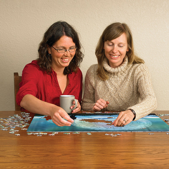 Two women enjoy assembling the 'Two Wolves' jigsaw puzzle from Cobble Hill at a wooden table. One woman holds a mug while both focus on placing puzzle pieces, surrounded by scattered pieces and a beautiful image of wolves in a serene landscape.