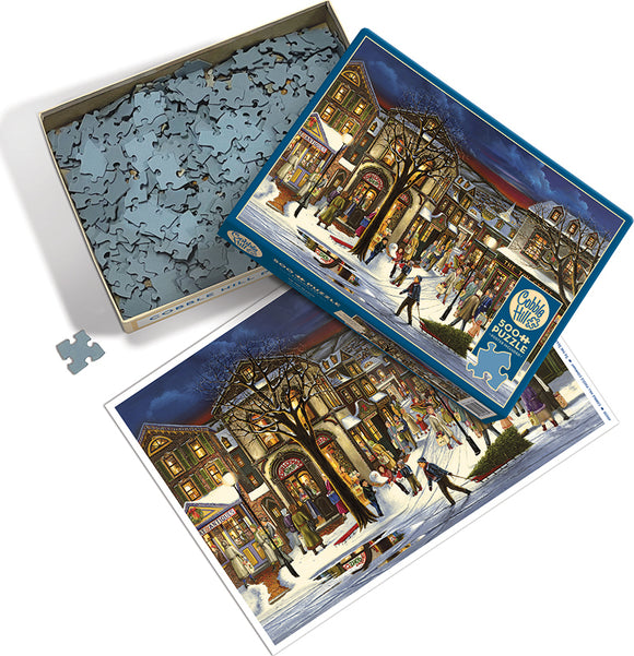 The product titled 'Tis the Season' by Cobble Hill is a jigsaw puzzle featuring a festive winter scene. The image shows an open box with numerous jigsaw pieces scattered around. The top lid displays a colorful illustration of a lively holiday street scene with people enjoying various winter activities, snow-covered buildings, and trees adorned with lights. The scene captures a cozy, charming atmosphere of the holiday season, perfect for family gatherings and seasonal fun.