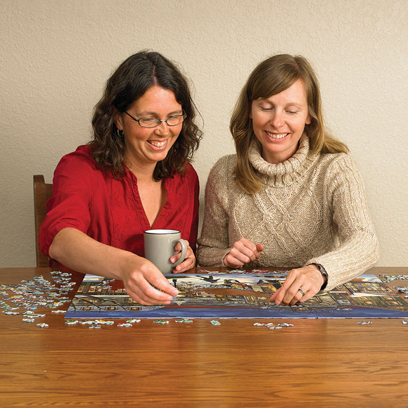 Two women engaged in putting together a jigsaw puzzle titled 'Tis the Season' by Cobble Hill. One woman, wearing a red shirt, is placing a piece while holding a grey mug. The other woman, dressed in a beige sweater, smiles as she helps. The puzzle features a festive winter scene, with colorful pieces spread out on a wooden table.