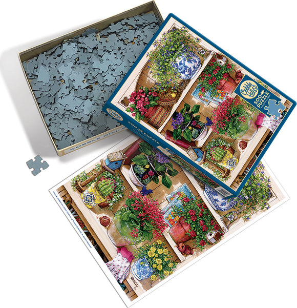 The Flower Cupboard jigsaw puzzle by Cobble Hill features a vibrant and intricately illustrated scene of a cupboard filled with colorful plants and flowers, displayed in pots and jars. The packaging includes a picture of the completed puzzle laid out beside the partially opened box, revealing the collection of puzzle pieces inside.