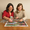 Two women sitting at a wooden table, joyfully working on the Flower Cupboard jigsaw puzzle by Cobble Hill. One woman holds a cup while placing a puzzle piece, and colorful puzzle pieces are scattered around them.