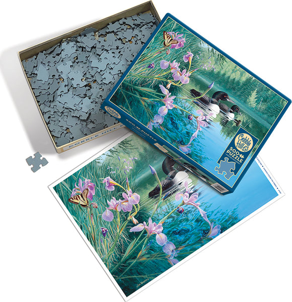 A glimpse of the Cobble Hill Iris Cove Loons jigsaw puzzle. The box features a vibrant image of loons on a serene lake surrounded by blooming irises and a butterfly. The puzzle pieces are packed in the box with some scattered around. The illustration showcases nature's beauty, making it perfect for puzzle enthusiasts who enjoy wildlife themes.