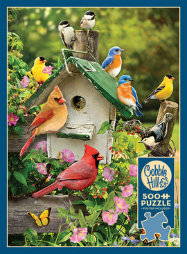 A colorful jigsaw puzzle titled 'Summer Birdhouse' by Cobble Hill, featuring a beautifully illustrated scene of various birds, including a cardinal, bluebird, and yellow finches, gathered around a rustic birdhouse adorned with blooming flowers and greenery. The puzzle contains 500 pieces and includes a poster of the image.