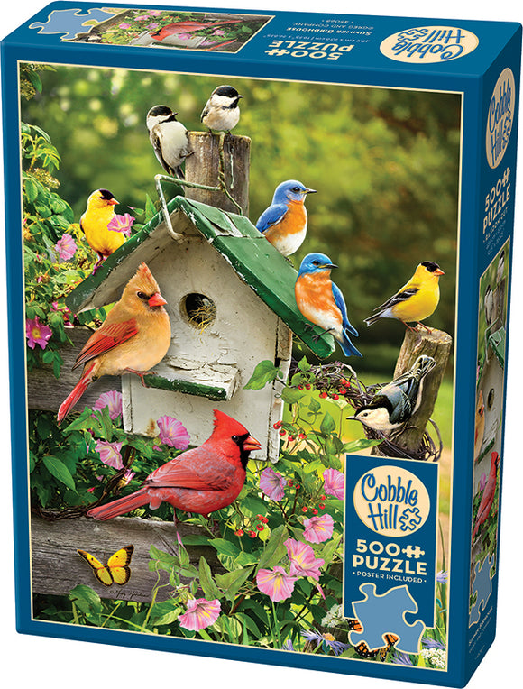 Cobble Hill 500-piece jigsaw puzzle titled 'Summer Birdhouse'. The box features a vibrant scene of a wooden birdhouse surrounded by colorful flowers and various birds, including a cardinal and bluebird, set against a lush green background. A butterfly can also be seen, adding to the enchanting wildlife theme.
