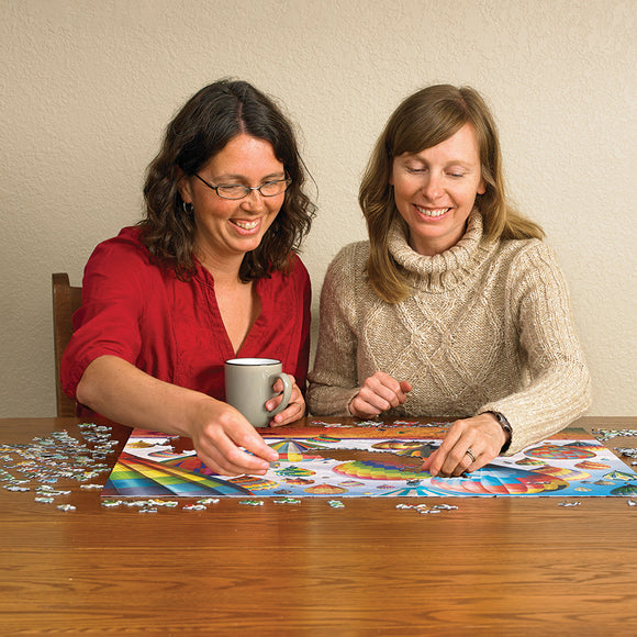 Two women smiling while assembling the 'Up in the Air' jigsaw puzzle by Cobble Hill. One woman holds a mug, and they work on a colorful puzzle featuring hot air balloons on a wooden table.