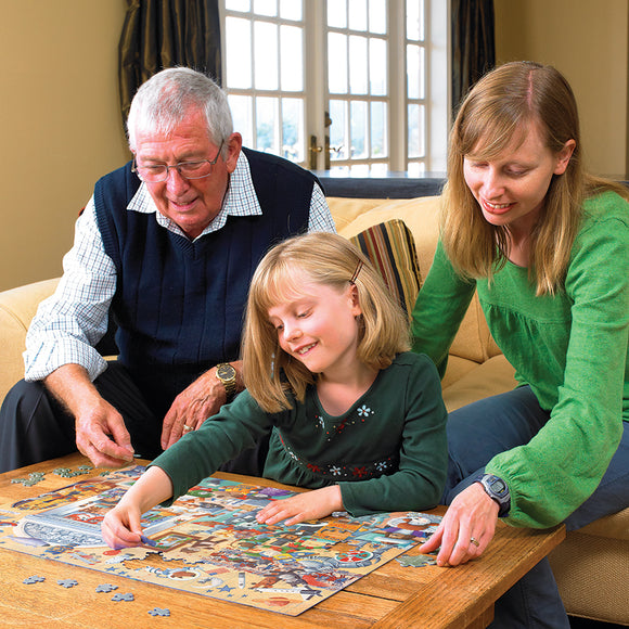 A family enjoying a jigsaw puzzle titled 'Cats and Dogs Museum' by Cobble Hill. A grandfather, daughter, and mother are engaged in assembling colorful pieces together on a wooden table in a cozy living room.
