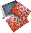 A colorful jigsaw puzzle titled 'Springtime Goldfinches' by Cobble Hill. The image showcases bright illustrations of goldfinches perched amidst vibrant flowers and foliage. The puzzle pieces are spread out in an open box, with a reference image of the completed puzzle alongside, capturing the rich colors and intricate details of the design.
